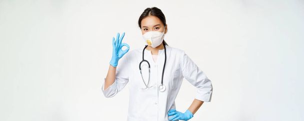 Sonriendo médico asiático, médico femenino muestra signo de bien, lleva respirador de la captura de la enfermedad covid, de pie en uniforme sobre fondo blanco. - Foto, Imagen