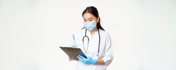 Asiatische Ärztin schreibt Patientendaten auf Klemmbrett, schreibt Rezept oder Diagnose, steht in medizinischer Maske mit Gummihandschuhen, weißer Hintergrund. - Foto, Bild