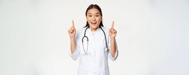 Excited smiling nurse, female doctor in medical uniform pointing up, showing promo sale, healthcare clinic discount, standing over white background. - Фото, изображение
