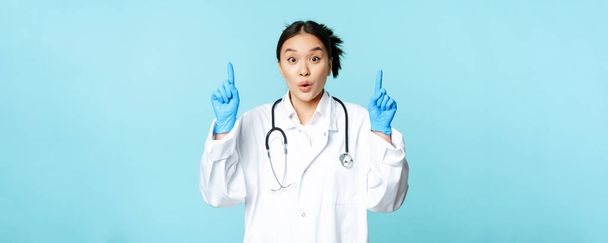 Ενθουσιασμένη Ασιάτισσα ιατρός, γυναίκα γιατρός που δείχνει τα δάχτυλα ψηλά, φοράει αποστειρωμένα γάντια και στολή, στέκεται πάνω από το μπλε φόντο. - Φωτογραφία, εικόνα