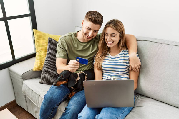 Jeune couple hispanique souriant heureux en utilisant ordinateur portable et carte de crédit assis sur le canapé avec chien à la maison. - Photo, image