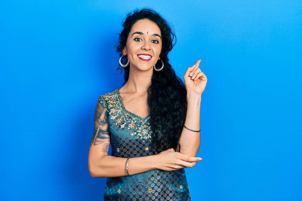 Young woman wearing bindi and traditional kurta dress with a big smile on face, pointing with hand and finger to the side looking at the camera.  - Photo, Image