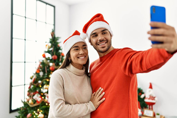 Νεαρό ζευγάρι ισπανόφωνων που φορούν χριστουγεννιάτικο καπέλο κάνοντας selfie από το smartphone στο σπίτι. - Φωτογραφία, εικόνα