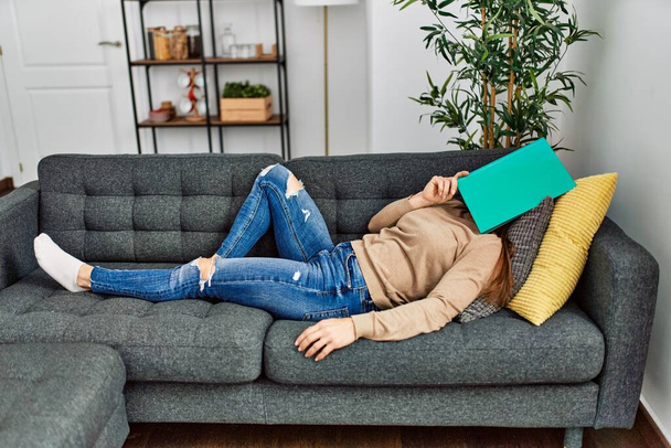 Καυκάσια γυναίκα μέσης ηλικίας κοιμάται στον καναπέ που καλύπτει το πρόσωπο με βιβλίο στο σπίτι - Φωτογραφία, εικόνα
