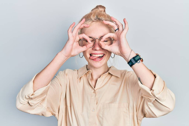 Beautiful caucasian woman with blond hair wearing casual look and glasses doing ok gesture like binoculars sticking tongue out, eyes looking through fingers. crazy expression.  - Foto, Imagem