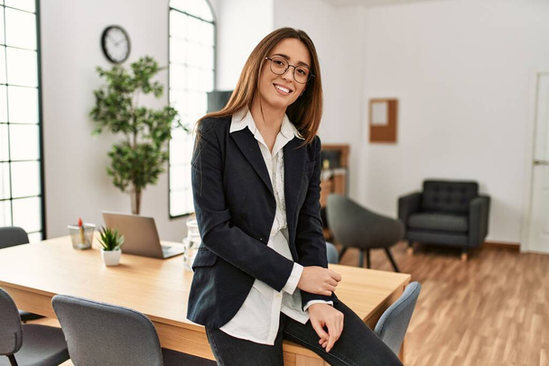 Young hispanic woman business worker smiling confident sitting on table at office - Fotó, kép