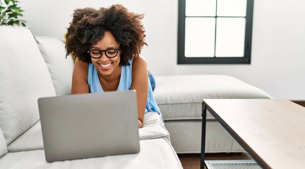 Νεαρή Αφροαμερικανή γυναίκα χαμογελά με αυτοπεποίθηση χρησιμοποιώντας φορητό υπολογιστή στο σπίτι - Φωτογραφία, εικόνα