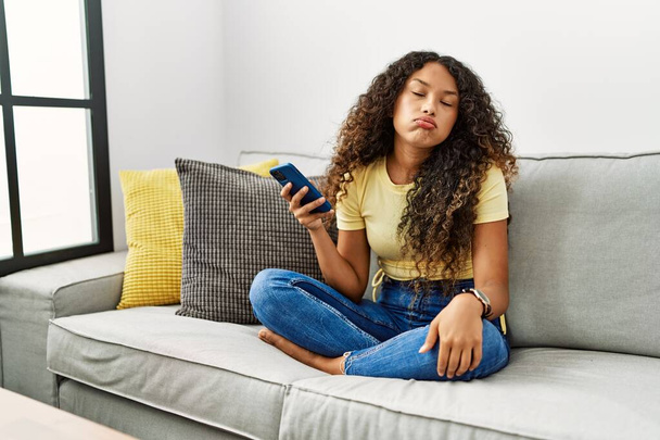 Beautiful hispanic woman sitting on the sofa at home using smartphone looking sleepy and tired, exhausted for fatigue and hangover, lazy eyes in the morning.  - Photo, Image