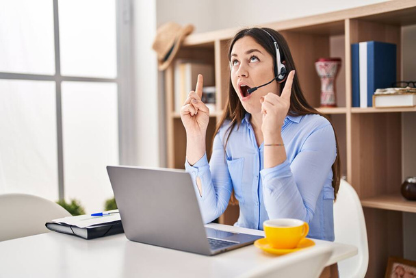 Young brunette woman wearing call center agent headset amazed and surprised looking up and pointing with fingers and raised arms.  - Zdjęcie, obraz
