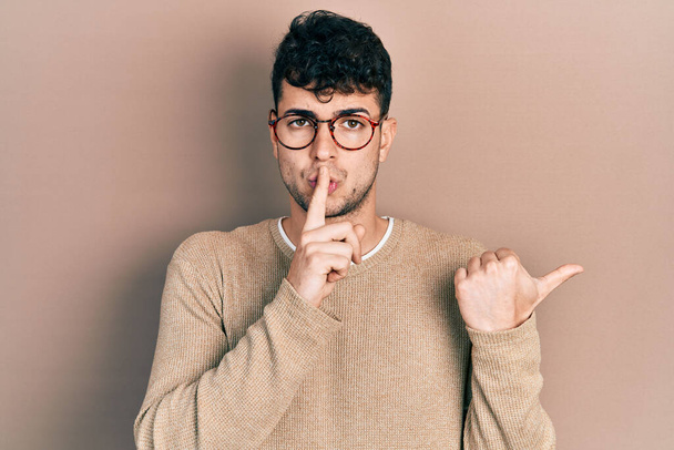 Mladý hispánec v neformálním oblečení a brýlích žádá, aby byl zticha s prstem na rtech a ukazoval rukou na stranu. mlčení a tajný koncept.  - Fotografie, Obrázek