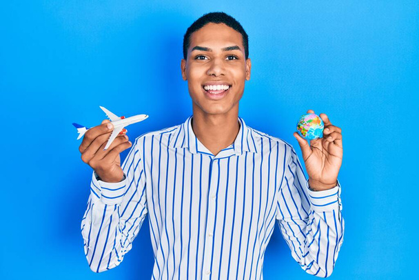 Молодой африканский американец держит бумажный самолет и мировой шар улыбается с счастливой и прохладной улыбкой на лице. показывая зубы.  - Фото, изображение