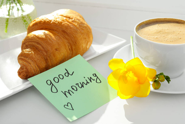 Cup of aromatic coffee with croissant, beautiful yellow freesia and Good Morning note on windowsill - Photo, Image
