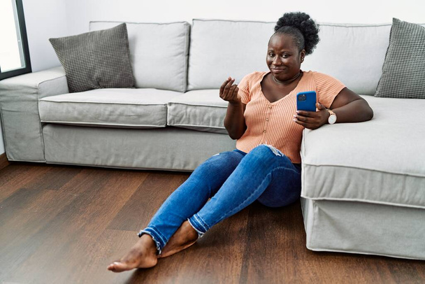 Mujer africana joven usando teléfono inteligente sentado en el suelo en casa haciendo gesto de dinero con las manos, pidiendo pago de sueldo, negocio millonario  - Foto, imagen