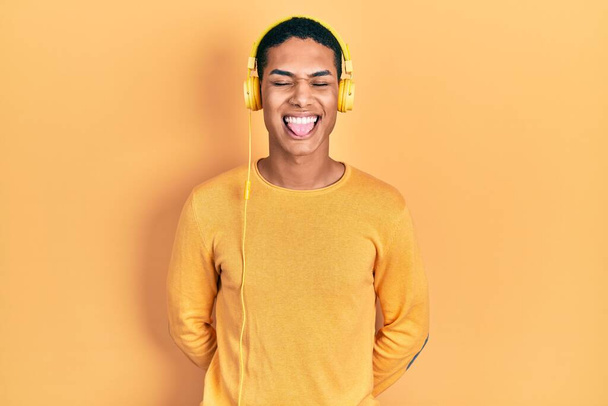 Молодой африканский американец слушает музыку, используя наушники, торчащие языком, довольный смешным выражением лица. концепция эмоций.  - Фото, изображение