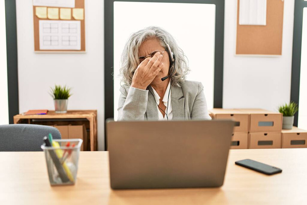 Femme d'affaires d'âge moyen assis sur le bureau travaillant à l'aide d'un ordinateur portable au bureau fatigué se frottant le nez et les yeux ressentant fatigue et maux de tête. concept de stress et de frustration.  - Photo, image