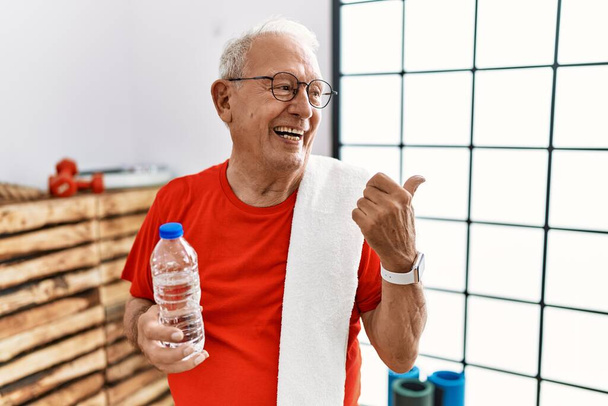 Homme âgé portant des vêtements de sport et une serviette à la salle de gym souriant avec le visage heureux regardant et pointant vers le côté avec le pouce vers le haut.  - Photo, image
