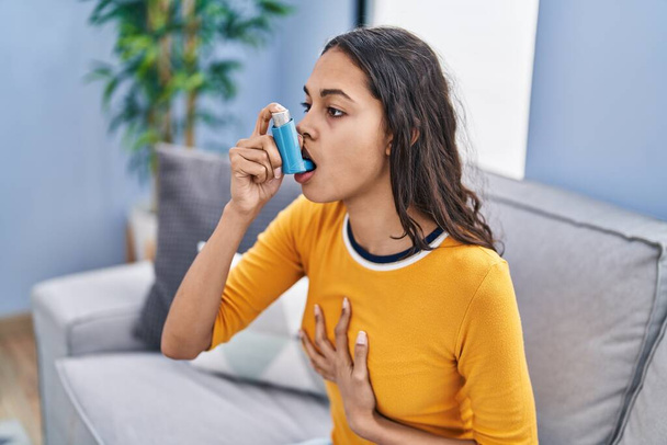 Young african american woman using inhaler sitting on sofa at home - Foto, Bild