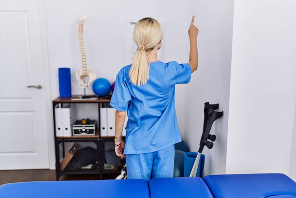 Young physiotherapist woman working at pain recovery clinic posing backwards pointing ahead with finger hand  - Valokuva, kuva