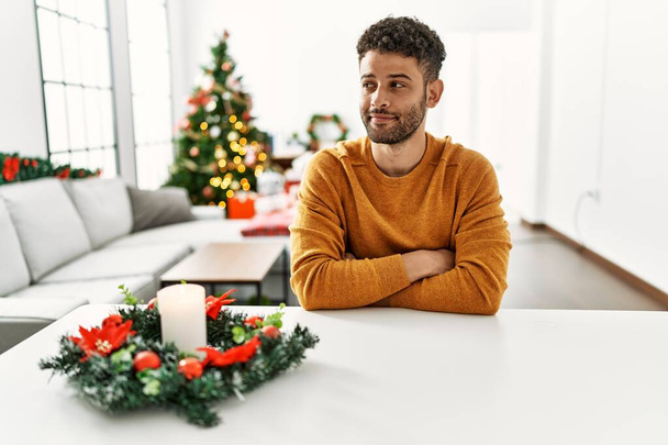 Arabischer junger Mann sitzt auf dem Tisch am Weihnachtsbaum, lächelt zur Seite und starrt nachdenklich weg.  - Foto, Bild