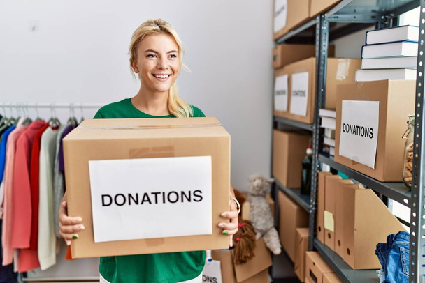 Młoda blondynka wolontariuszka uśmiecha się szczęśliwa trzymając pudełko z datkami w centrum charytatywnym. - Zdjęcie, obraz