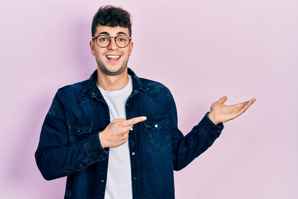 Νεαρός Ισπανός άνδρας φορώντας καθημερινά ρούχα και γυαλιά κατάπληκτος και χαμογελώντας στην κάμερα ενώ παρουσιάζει με το χέρι και δείχνοντας με το δάχτυλο.  - Φωτογραφία, εικόνα