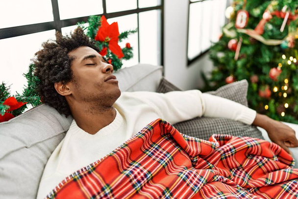 Νεαρός Αφροαμερικάνος κοιμάται ξαπλωμένος στον καναπέ δίπλα στο χριστουγεννιάτικο δέντρο στο σπίτι. - Φωτογραφία, εικόνα
