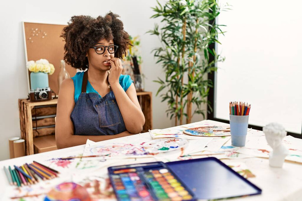 Gyönyörű afro-amerikai nő afro hajfestés a művészeti stúdióban úgy néz ki, stresszes és ideges a kezét a száj harapdáló körmök. szorongás.  - Fotó, kép