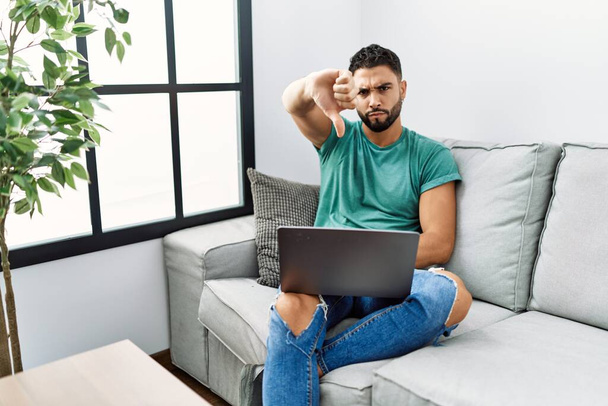 Young handsome man with beard using computer laptop sitting on the sofa at home looking unhappy and angry showing rejection and negative with thumbs down gesture. bad expression.  - Fotografie, Obrázek