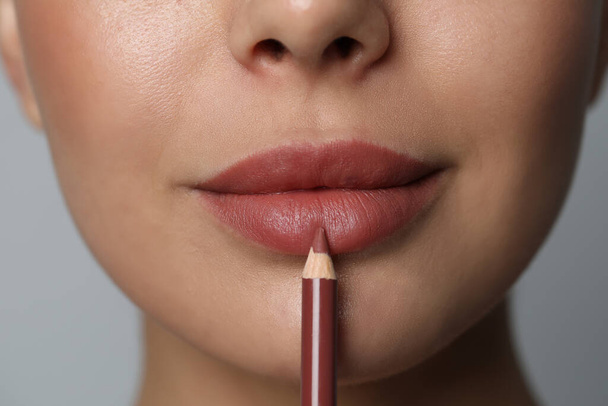 Young woman applying beautiful nude lip pencil on grey background, closeup - Fotó, kép