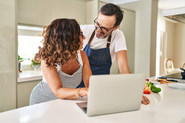 Orta yaşlı İspanyol çift mutfakta yemek pişiriyor ve laptop kullanıyor. - Fotoğraf, Görsel