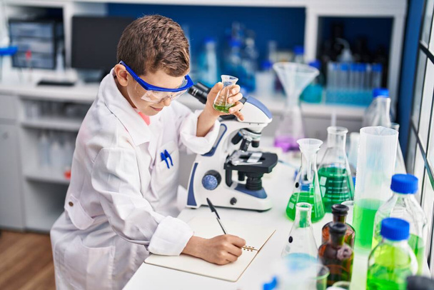 Blond child wearing scientist uniform writing on notebook at laboratory - Фото, изображение