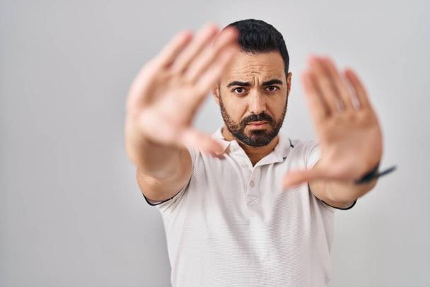 Jeune homme hispanique avec barbe portant des vêtements décontractés sur fond blanc faisant cadre en utilisant les mains paumes et les doigts, perspective de la caméra  - Photo, image