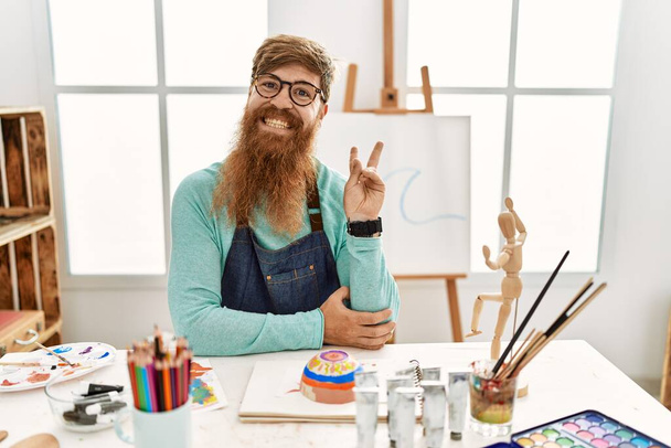 Vörös hajú férfi hosszú szakállal, agyagtálat festett a művészeti stúdióban, mosolyogva, boldog arccal kacsintott a kamerára, győzelmi jelet téve. Második..  - Fotó, kép