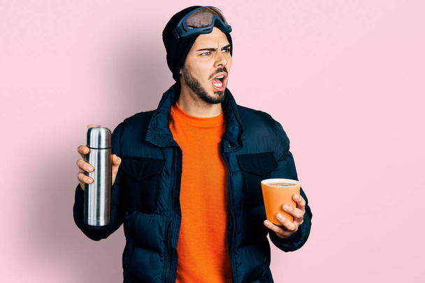 Young hispanic man with beard wearing snow wear and sky glasses holding coffee and thermo angry and mad screaming frustrated and furious, shouting with anger. rage and aggressive concept.  - Photo, Image