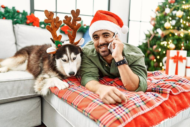 Junger hispanischer Mann spricht auf dem Smartphone liegend auf Sofa mit Hund am Weihnachtsbaum zu Hause - Foto, Bild