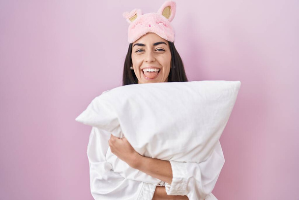Молода брюнетка жінка в масці для сну і піжамі обіймає подушку, що стирчить язиком щасливо зі смішним виразом
.  - Фото, зображення