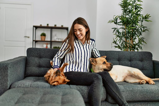 Jeune femme hispanique souriante confiante assise sur un canapé avec des chiens à la maison - Photo, image