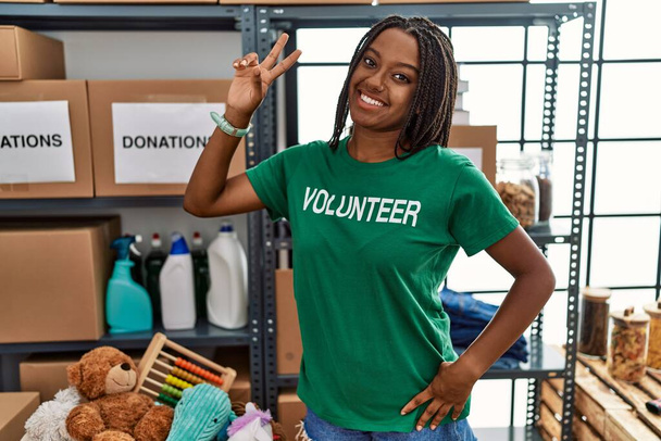 Молодая африканская американка, работающая в футболке волонтера на пожертвованиях, улыбается, глядя в камеру, показывая пальцы, делающие знак победы. Номер два.  - Фото, изображение