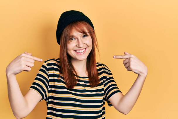 Κοκκινομάλλα νεαρή γυναίκα φορώντας μάλλινο καπέλο που δείχνει σίγουρος με χαμόγελο στο πρόσωπο, δείχνοντας τον εαυτό του με τα δάχτυλα περήφανος και χαρούμενος.  - Φωτογραφία, εικόνα