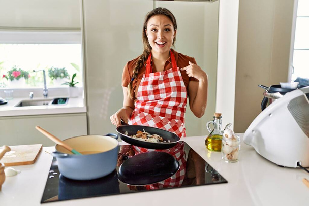 Mutfakta önlük giymiş genç, güzel esmer bir kadın, mutlu ve gururlu bir gülümsemeyi işaret ediyor.  - Fotoğraf, Görsel