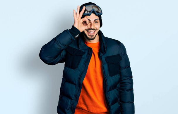 Young hispanic man with beard wearing snow wear and sky glasses doing ok gesture with hand smiling, eye looking through fingers with happy face.  - Fotoğraf, Görsel