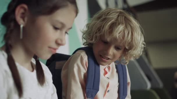 Two cute schoolchildren playing together in school corridor close up. Portrait of focused girl look down sitting hallway with smiling curly blond schoolboy. Curious preteens kids have fun on break. - Footage, Video