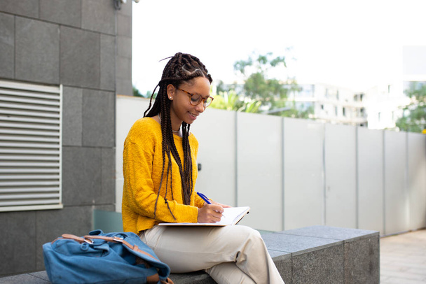 Retrato de estudante africana sentada do lado de fora estudando com caneta e livro - Foto, Imagem