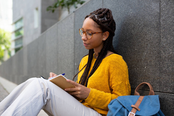 Πορτρέτο της νεαρής Αφροαμερικανής φοιτήτριας που κάθεται έξω και γράφει στο βιβλίο - Φωτογραφία, εικόνα