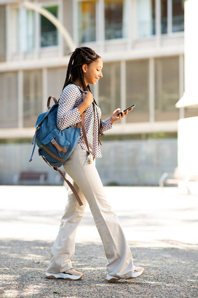 Pełna długość boczny portret młodej Afryki amerykańskiej studentka spaceru z torbą smsowania z jej telefon komórkowy na zewnątrz w mieście - Zdjęcie, obraz