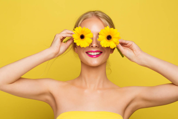 Woman covering Eyes with Yellow Flowers. Happy Smiling Model with Gerbera Floral Sun Glasses over isolated Orange background. Cheerful Summer Fashion Girl - 写真・画像