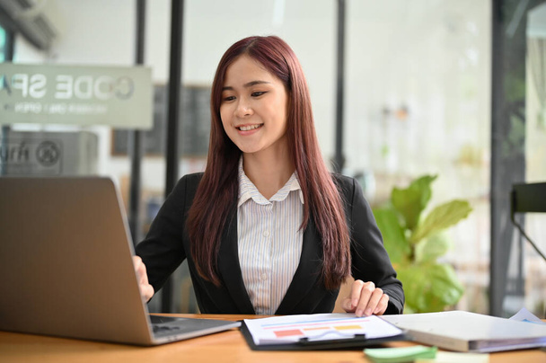 Attractive young asian businesswoman of female office employee sits at her office desk, working on her financial paperwork and using laptop computer. - Photo, Image