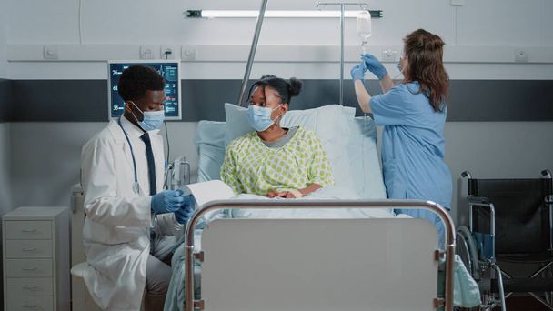 Kranker Patient, der auf der Krankenhausstation von einem Arzt mit Mundschutz medizinisch versorgt wird. Arzt spricht mit Frau, während Assistentin bei Pandemie mit IV-Tropfsack hilft. - Foto, Bild
