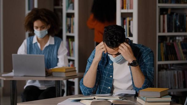 Aşırı yorgun, koruyucu maskeli genç Arap erkek öğrenci kütüphanede oturuyor ödev hazırlıyor sınava hazırlanıyor, hata yapıyor elle başını kavrıyor şok krizi geçiriyor. - Fotoğraf, Görsel