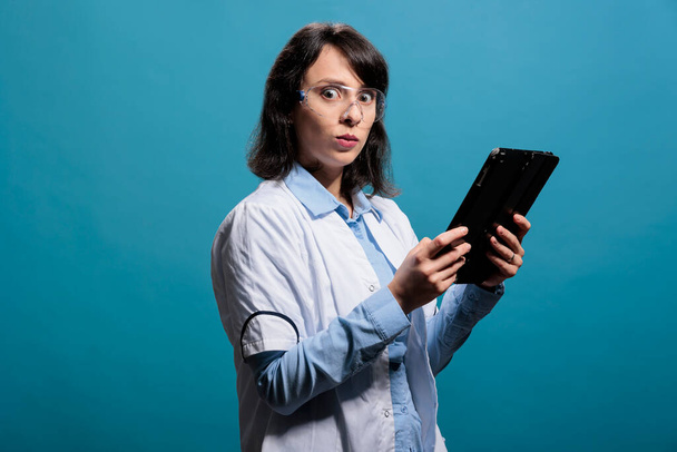 Chemiker mit modernem Touchscreen-Tablet, Schutzbrille und Laborkittel auf blauem Hintergrund. Junge erwachsene Biologie-Forscherin mit elektronischem Gerät auf blauem Hintergrund. - Foto, Bild